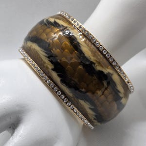 May include: A wide bangle bracelet featuring a snakeskin design in brown, tan, and black. The bracelet has a gold-tone trim and is embellished with rows of small, clear crystals. The jewellery is displayed on a white hand.