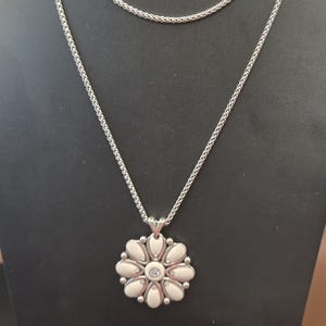 Puede incluir: Un collar plateado con un colgante de flor. El colgante presenta una piedra central transparente rodeada de elementos blancos en forma de p&eacute;talo. El collar tiene un dise&ntilde;o de cadena delicado y se exhibe en un maniqu&iacute; negro.