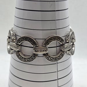 May include: Silver-toned bracelet with an ornate design. The bracelet features oval-shaped links with detailed scrollwork and connecting bars. The bracelet is displayed on a white, cone-shaped object with black horizontal lines.