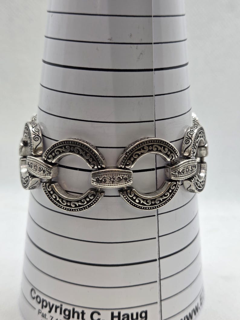 May include: Silver-toned bracelet with an ornate design. The bracelet features oval-shaped links with detailed scrollwork and connecting bars. The bracelet is displayed on a white, cone-shaped object with black horizontal lines.