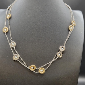 May include: A silver necklace with two strands, featuring alternating gold-toned and silver-toned circular pendants. Each pendant holds a small, round, white pearl. The necklace is displayed on a black mannequin.
