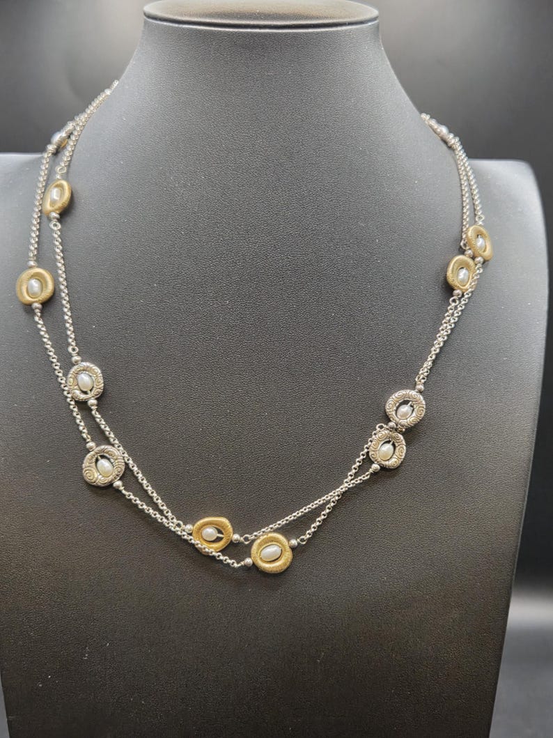May include: A silver necklace with two strands, featuring alternating gold-toned and silver-toned circular pendants. Each pendant holds a small, round, white pearl. The necklace is displayed on a black mannequin.