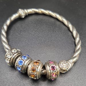 Puede incluir: Pulsera plateada con dise&ntilde;o de banda trenzada. Cuenta con cuentas decorativas con piedras preciosas azules, naranjas y rosas. La pulsera se muestra sobre una superficie oscura. El dise&ntilde;o es intrincado.
