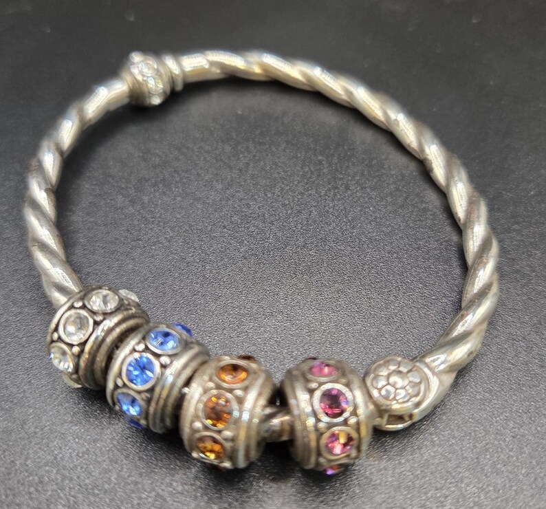 Puede incluir: Pulsera plateada con dise&ntilde;o de banda trenzada. Cuenta con cuentas decorativas con piedras preciosas azules, naranjas y rosas. La pulsera se muestra sobre una superficie oscura. El dise&ntilde;o es intrincado.