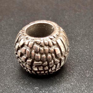 May include: A silver-colored bead with a textured surface. The bead has a cylindrical hole running through the center. The surface is covered in a pattern of raised, irregular shapes.