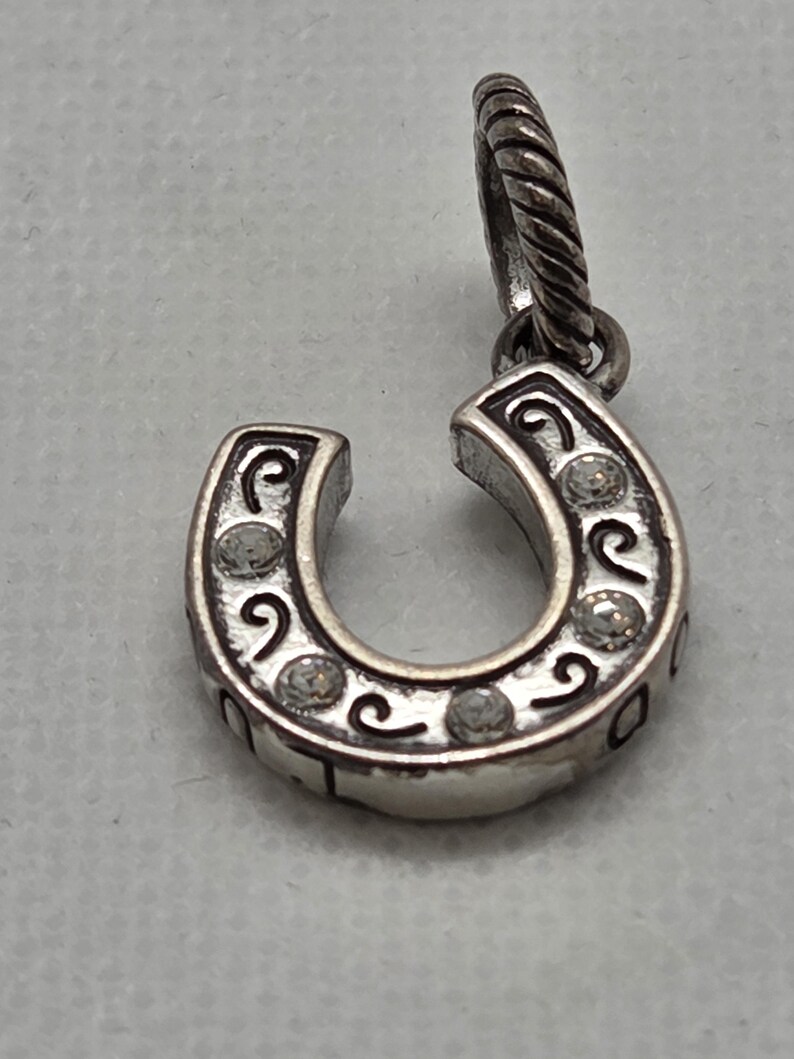 May include: Silver-toned horseshoe charm with clear crystal accents. The horseshoe is detailed with scrollwork and small, round crystals. The charm has a twisted bail for attaching to a necklace or bracelet. The horseshoe is a symbol of good luck.