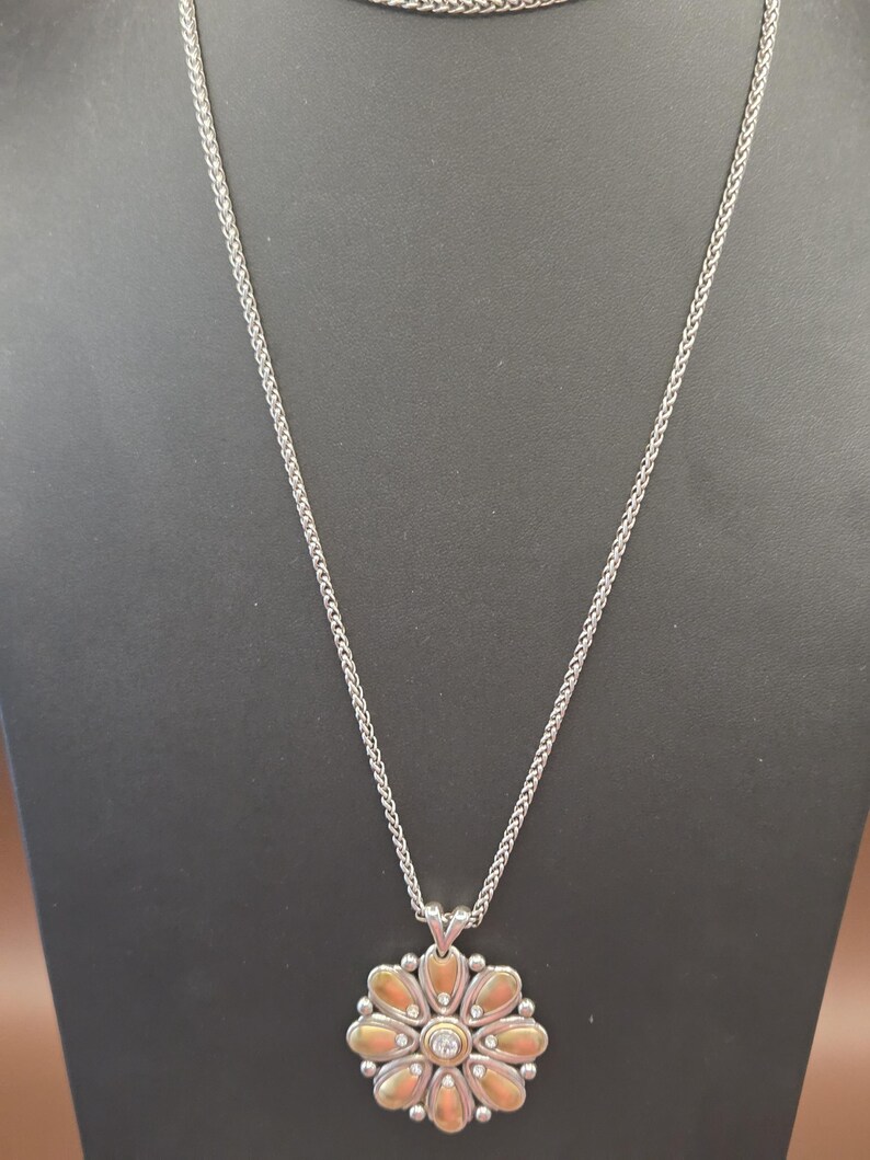 Puede incluir: Collar plateado con un colgante de flor. El colgante presenta una piedra central transparente rodeada de elementos en forma de p&eacute;talo en oro y plata. La cadena es de dise&ntilde;o delicado y tejido.