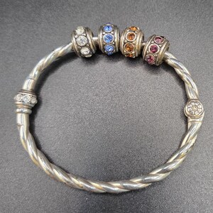 Puede incluir: Pulsera de brazalete plateada con un dise&ntilde;o trenzado. La pulsera presenta cuatro cuentas decorativas con piedras preciosas de colores: transparentes, azules, naranjas y rosas. La pulsera tiene un dise&ntilde;o floral en el cierre.