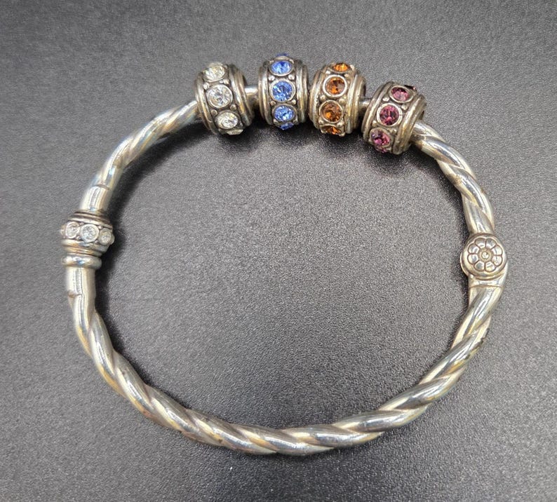 Puede incluir: Pulsera de brazalete plateada con un dise&ntilde;o trenzado. La pulsera presenta cuatro cuentas decorativas con piedras preciosas de colores: transparentes, azules, naranjas y rosas. La pulsera tiene un dise&ntilde;o floral en el cierre.