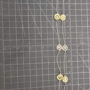 May include: A long silver necklace with alternating gold and silver-toned circular pendants. The necklace is displayed on a black cutting mat with grid lines and measurement markings.