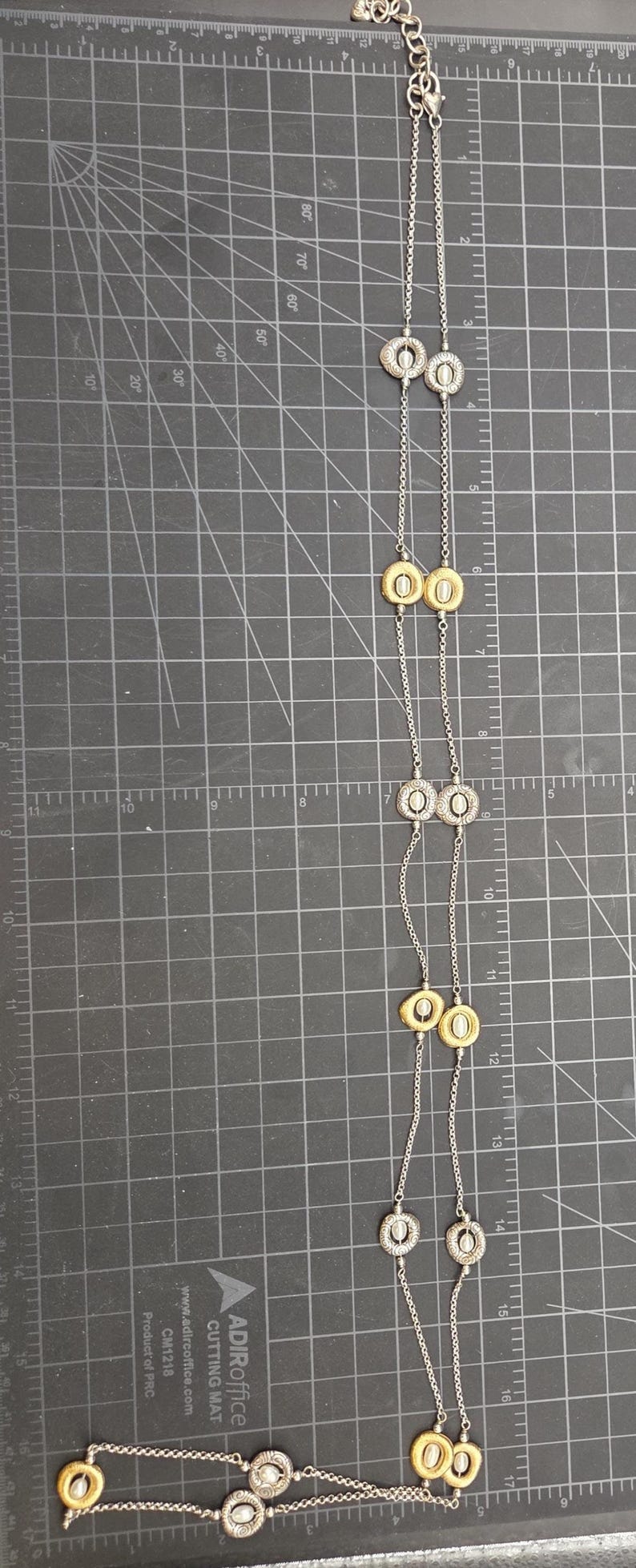 May include: A long silver necklace with alternating gold and silver-toned circular pendants. The necklace is displayed on a black cutting mat with grid lines and measurement markings.