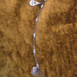 May include: Silver-toned bracelet with a heart-shaped charm and a small tag that says "MOM". The bracelet has a chain design with decorative beads and a lobster clasp closure. The heart charm has a floral design.