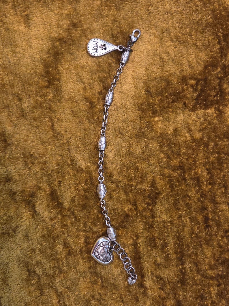 May include: Silver-toned bracelet with a heart-shaped charm and a small tag that says "MOM". The bracelet has a chain design with decorative beads and a lobster clasp closure. The heart charm has a floral design.