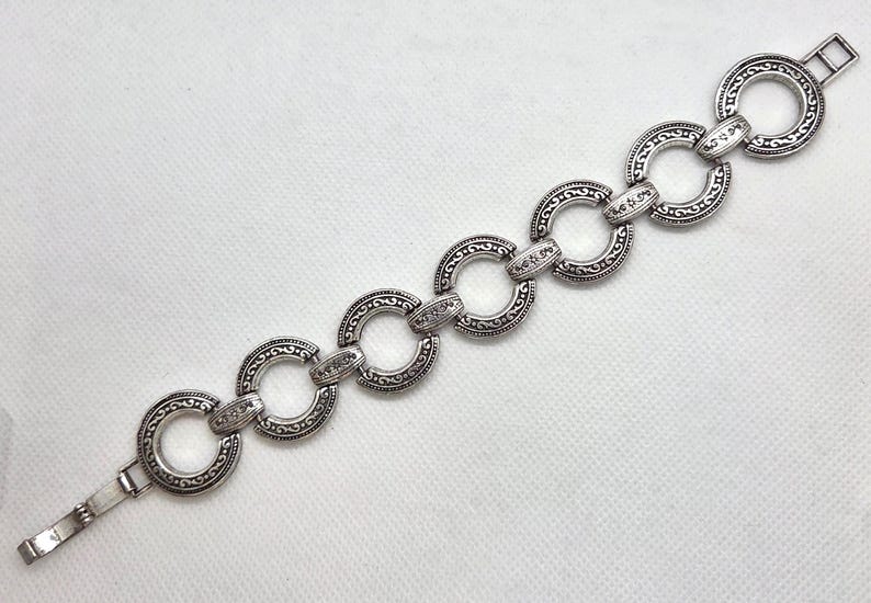 May include: Silver-toned bracelet featuring a series of interconnected, circular links. Each link is adorned with an intricate, embossed pattern and connected by smaller, decorative elements. The bracelet has a clasp closure.