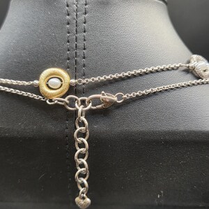 May include: A silver necklace with a gold pendant holding a small pearl. The necklace has a delicate chain design and a heart-shaped charm at the clasp. The necklace is displayed on a black mannequin.