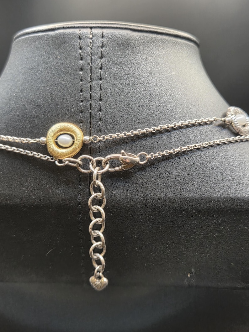 May include: A silver necklace with a gold pendant holding a small pearl. The necklace has a delicate chain design and a heart-shaped charm at the clasp. The necklace is displayed on a black mannequin.
