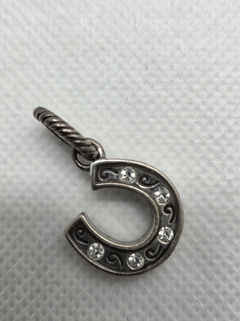 May include: Silver-toned horseshoe charm with clear crystal accents. The horseshoe is adorned with decorative swirls and five round crystals. The charm has a twisted loop for attachment to a necklace or bracelet.