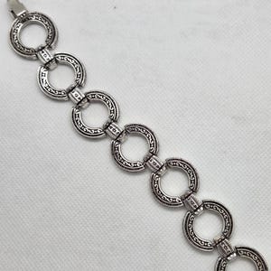 May include: Silver-toned bracelet featuring a series of interconnected, ornate circular links. Each link is detailed with a swirling, patterned design. The bracelet has a clasp closure.