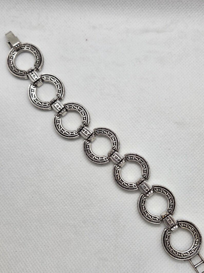 May include: Silver-toned bracelet featuring a series of interconnected, ornate circular links. Each link is detailed with a swirling, patterned design. The bracelet has a clasp closure.