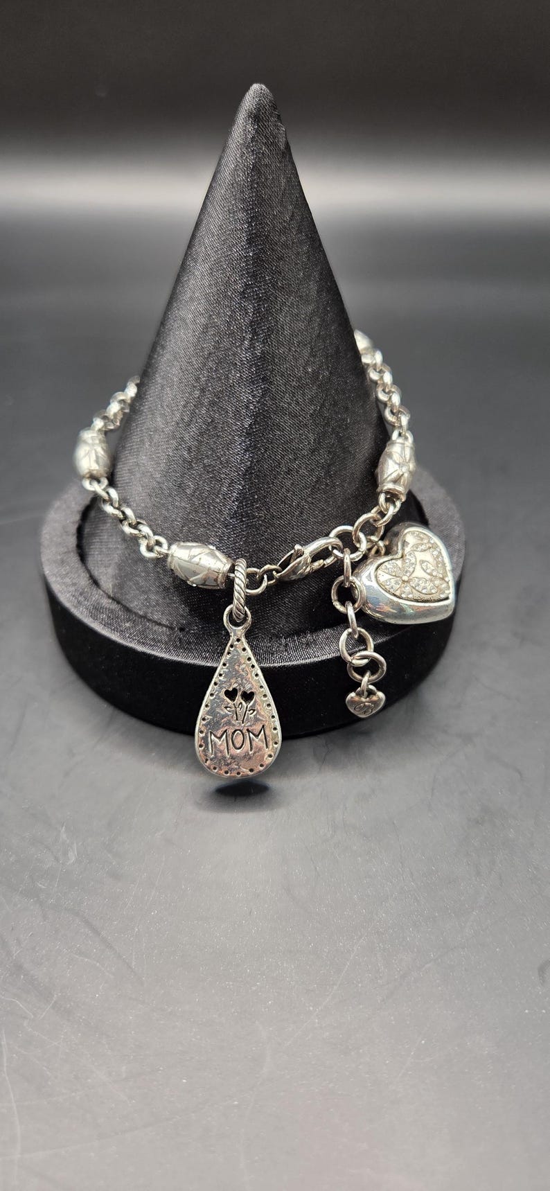 May include: Silver bracelet with a chain-link design, featuring decorative beads and charms. Charms include a heart with embedded crystals and a teardrop pendant with the word "MOM". The bracelet is displayed on a black stand.