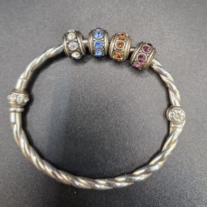 Puede incluir: Pulsera plateada con un dise&ntilde;o de banda trenzada. La pulsera presenta cuatro cuentas decorativas con piedras preciosas transparentes, azules, naranjas y moradas. Una joya.