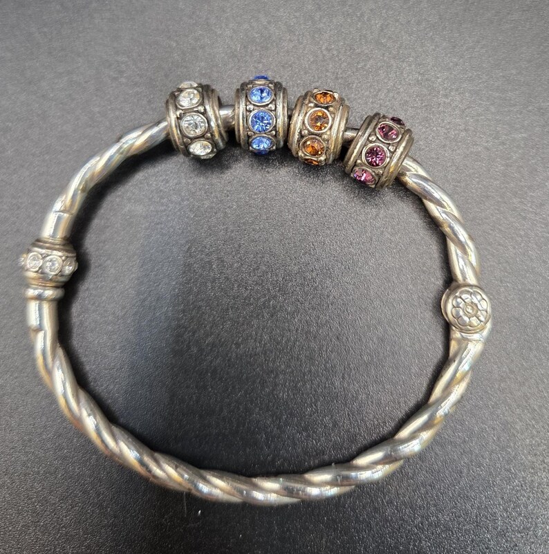 Puede incluir: Pulsera plateada con un dise&ntilde;o de banda trenzada. La pulsera presenta cuatro cuentas decorativas con piedras preciosas transparentes, azules, naranjas y moradas. Una joya.