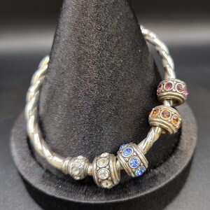 Puede incluir: Una pulsera plateada con un dise&ntilde;o retorcido. La pulsera presenta varias cuentas decorativas con piedras preciosas de colores, incluyendo cristales rosas, naranjas, azules y transparentes. La pulsera se exhibe en un soporte negro.