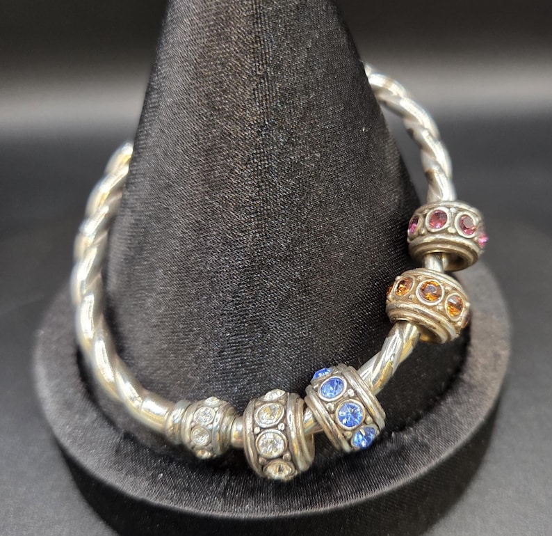 Puede incluir: Una pulsera plateada con un dise&ntilde;o retorcido. La pulsera presenta varias cuentas decorativas con piedras preciosas de colores, incluyendo cristales rosas, naranjas, azules y transparentes. La pulsera se exhibe en un soporte negro.
