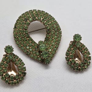 Pode incluir: Um conjunto de joias vintage com um broche e brincos a condizer. O broche tem formato de gota e os brincos têm formato de gota. Todas as peças são incrustadas com pequenas pedras de strass verde claro em metal dourado.