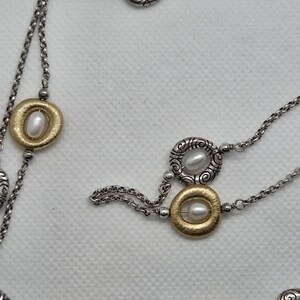 May include: A silver-coloured necklace with gold-coloured and silver-coloured circular accents, each featuring a pearl. The necklace has a delicate chain design. The jewellery is displayed against a white background.