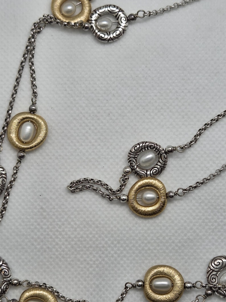 May include: A silver-coloured necklace with gold-coloured and silver-coloured circular accents, each featuring a pearl. The necklace has a delicate chain design. The jewellery is displayed against a white background.