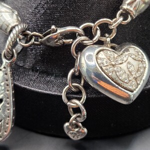 May include: Silver charm bracelet with a heart-shaped charm and a teardrop charm that says "MOM". The heart charm is decorated with small, clear stones. The bracelet is displayed on a black surface.