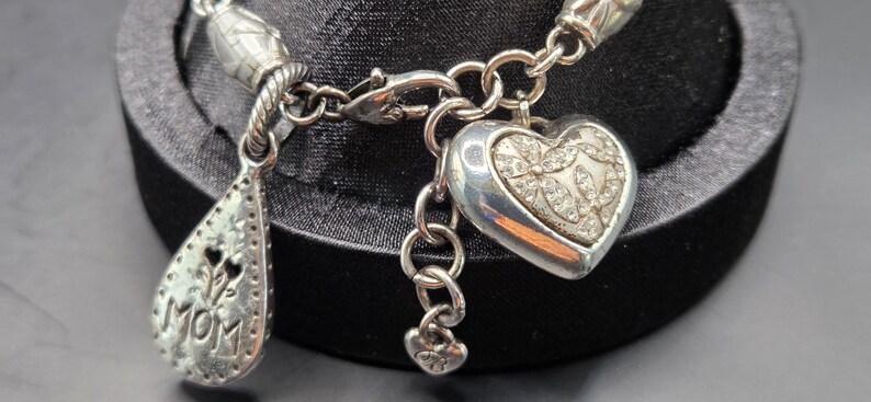 May include: Silver charm bracelet with a heart-shaped charm and a teardrop charm that says "MOM". The heart charm is decorated with small, clear stones. The bracelet is displayed on a black surface.