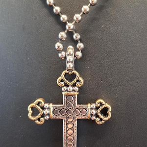 以下が含まれることがあります： 金色のアクセントが付いた、銀色のクロス ペンダント ネックレス。クロスは、円形とハート型の要素を備えた詳細なデザインです。ネックレスはボールチェーンです。