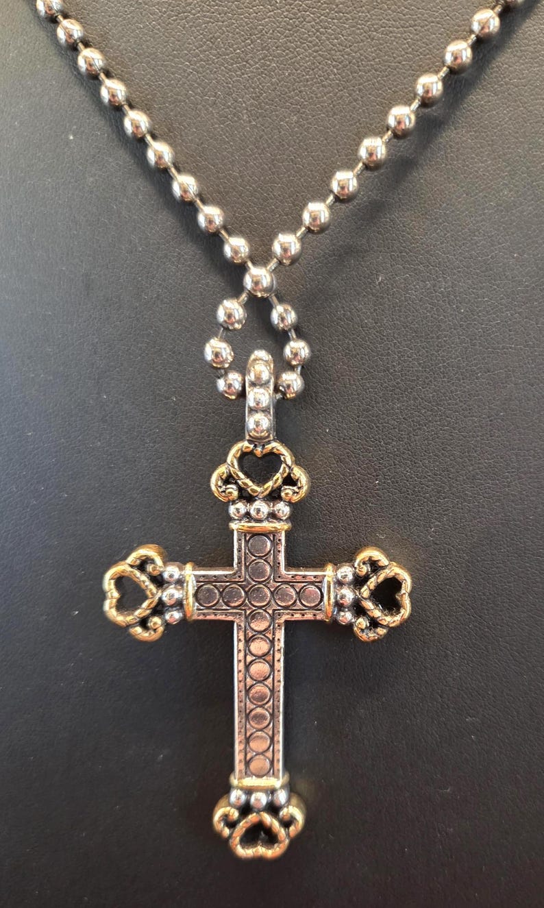 以下が含まれることがあります： 金色のアクセントが付いた、銀色のクロス ペンダント ネックレス。クロスは、円形とハート型の要素を備えた詳細なデザインです。ネックレスはボールチェーンです。