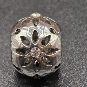 May include: Silver-toned bead with a floral design and a hammered texture. The bead features clear, round gemstones at the center and top. The bead has cut-out petal shapes around the sides.