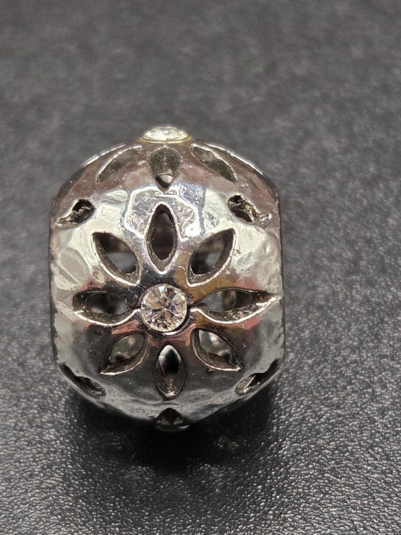 May include: Silver-toned bead with a floral design and a hammered texture. The bead features clear, round gemstones at the center and top. The bead has cut-out petal shapes around the sides.