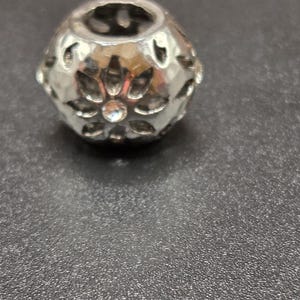 May include: A silver-colored bead with a floral design. The bead has a hollow center and is decorated with cut-out petal shapes and a central clear crystal. The bead is on a dark gray surface.