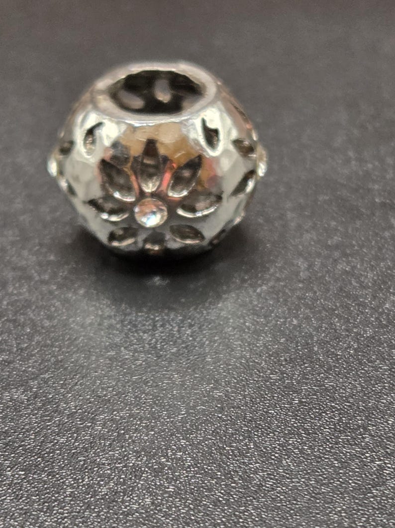 May include: A silver-colored bead with a floral design. The bead has a hollow center and is decorated with cut-out petal shapes and a central clear crystal. The bead is on a dark gray surface.