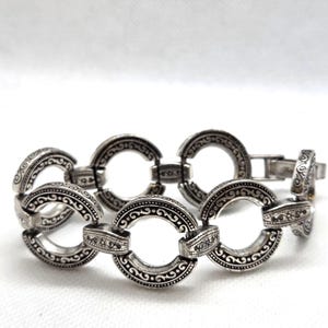 May include: Silver-toned bracelet featuring a series of interconnected, open circular links. Each link is adorned with intricate, swirling patterns and decorative detailing. The bracelet has a clasp closure and is displayed against a white background.