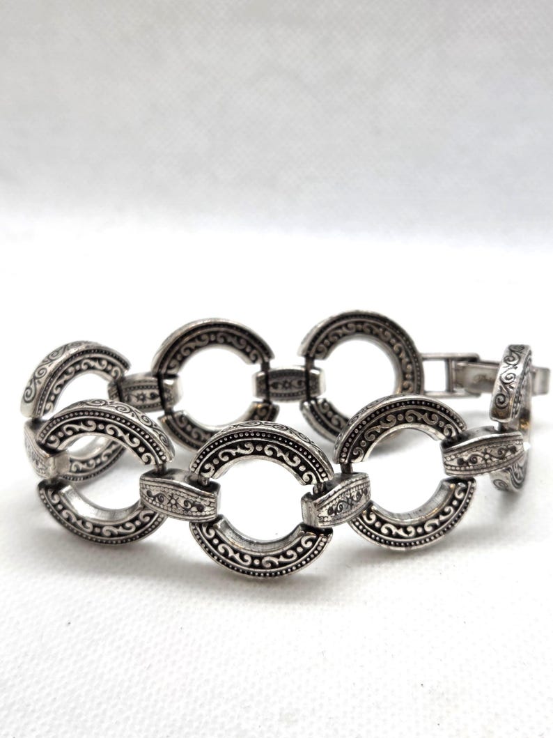 May include: Silver-toned bracelet featuring a series of interconnected, open circular links. Each link is adorned with intricate, swirling patterns and decorative detailing. The bracelet has a clasp closure and is displayed against a white background.