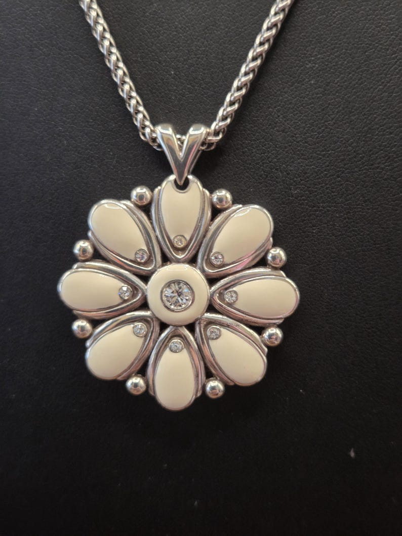 Puede incluir: Collar con colgante de flor plateado. El colgante presenta un dise&ntilde;o de flor esmaltada color crema con detalles de cristal. El collar tiene una cadena trenzada y un cierre de mosquet&oacute;n.