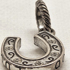 May include: Silver-toned horseshoe charm with clear crystal accents and decorative swirls. The charm has a twisted loop for attachment. The horseshoe is a symbol of good luck and is often used in jewelry.