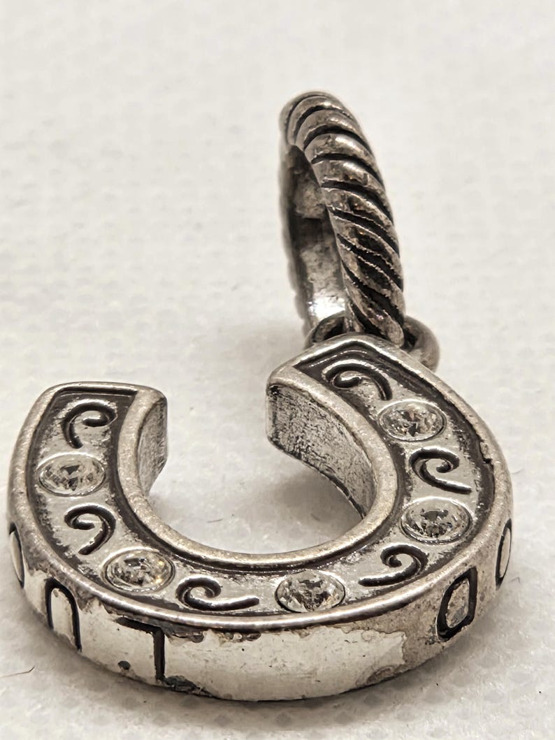 May include: Silver-toned horseshoe charm with clear crystal accents and decorative swirls. The charm has a twisted loop for attachment. The horseshoe is a symbol of good luck and is often used in jewelry.