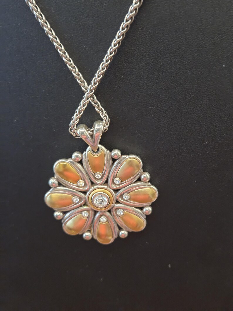 Puede incluir: Collar plateado con colgante de flor. El colgante presenta p&eacute;talos dorados, peque&ntilde;as piedras preciosas transparentes y una piedra preciosa central transparente. La cadena es de dise&ntilde;o tejido.