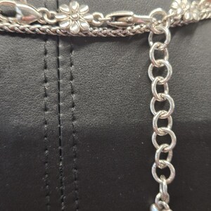 Puede incluir: Primer plano de un collar plateado con un cierre en forma de flor y un dije en forma de coraz&oacute;n. El collar tiene m&uacute;ltiples hilos y una cadena ajustable. El fondo es una superficie negra.