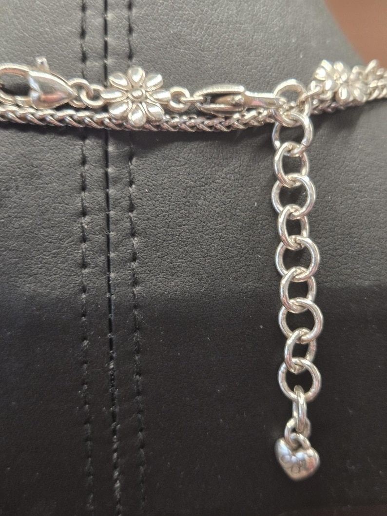 Puede incluir: Primer plano de un collar plateado con un cierre en forma de flor y un dije en forma de coraz&oacute;n. El collar tiene m&uacute;ltiples hilos y una cadena ajustable. El fondo es una superficie negra.