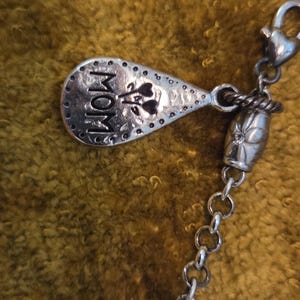 May include: A silver-toned charm with the word "MOM" and floral accents. The charm is attached to a chain with a decorative bead and a clasp. The background is a textured, golden-brown fabric.