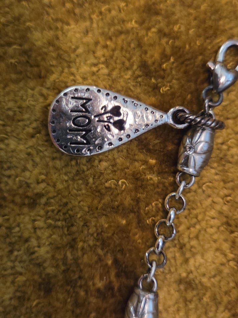 May include: A silver-toned charm with the word "MOM" and floral accents. The charm is attached to a chain with a decorative bead and a clasp. The background is a textured, golden-brown fabric.