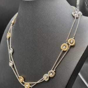 May include: A silver necklace with two strands, featuring gold and silver circular pendants with small, round, white pearls. The necklace is displayed on a black mannequin, highlighting its design and details.